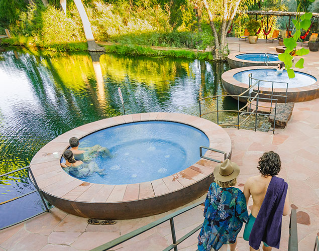 people in and walking towards 3 hot tub pools seated next to pond with trees surrounding it