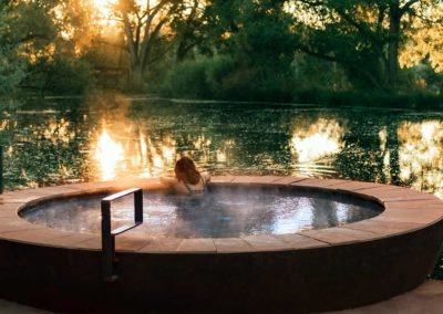 Woman in large hot tub overlooking pond and sun shining through tall trees