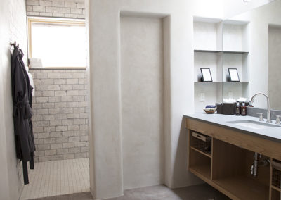 White bathroom with wood vanity and large mirror opposite a tiled walk in shower and robes hanging on the wall