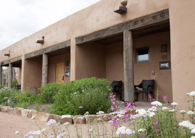 Front exterior of adobe building with desert landscaping and flowers in front of it and two chairs on the porch