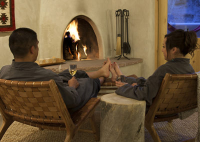 Couple wearing signature Ojo bathrobes drinking white wine in their room sitting on chairs in front of a kiva style fireplace