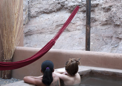 Two women soaking in private hot tub facing a red hammock with a mountain wall behind it