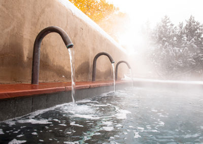 An outdoor wall with three faucets all pouring water into a thermal pool with snow on the trees in the background