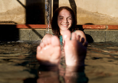 Woman with feet up floating in outdoor thermal pool with her head next to a spout and wall behind her