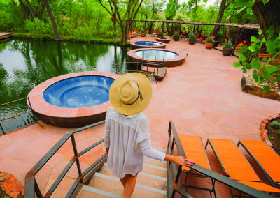 Woman in hat walking toward hot tub pools overlooking pond with lush trees in the background