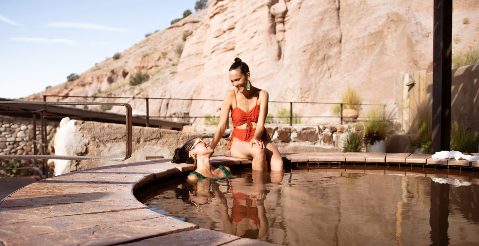 Ojo Caliente-Resort_0002_terrace pool duo Two people enjoying a round, outdoor hot spring pool. One is sitting on the edge wearing a red swimsuit, while the other is in the water, leaning on the edge in a green swimsuit. Rocky cliffs form the backdrop under a clear sky.