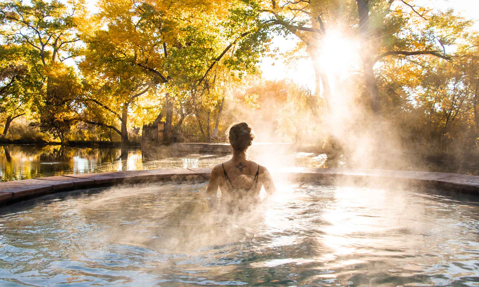 OSF_WebHero_Fall_Ojo-Santa-Fe-Soak A person with a tattoo on their upper back is sitting in a circular hot spring. Steam rises from the water. The hot spring is surrounded by autumn trees with golden leaves, and sunlight filters through the foliage.