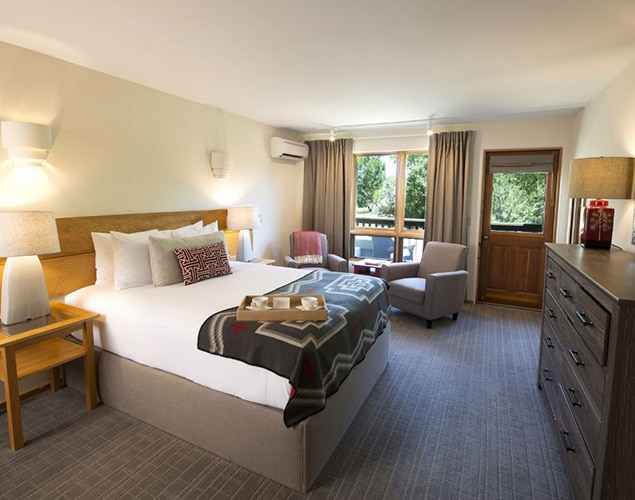 Queen Bed With Throw Blanket In Hotel Room With Seating Area By Window Overlooking Garden