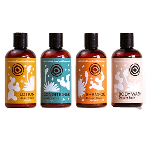 Four brown bottles labeled "Desert Rain" containing lotion, conditioner, shampoo, and body wash. The products are arranged from left to right and feature illustrations related to their contents. The labels also indicate they are made from local botanicals.