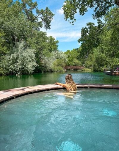 A person with long hair relaxes in a circular pool, partially submerged and facing away, overlooking a lush, green, tree-lined river under a sunny blue sky. The surroundings are serene with dense foliage and calm waters.