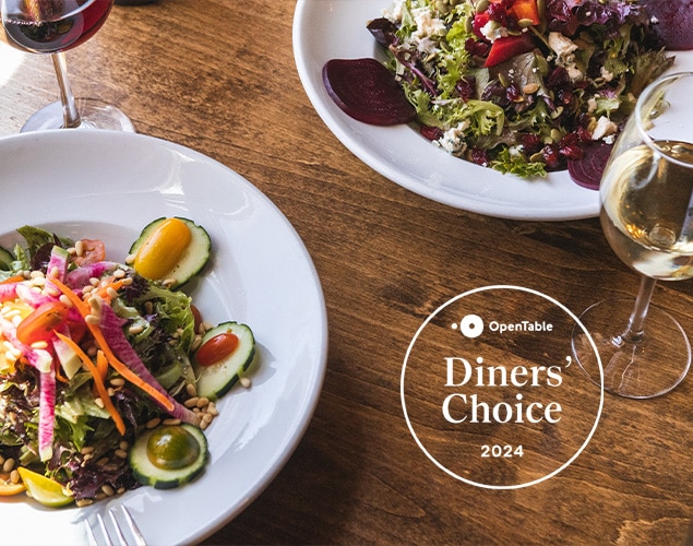 Two plates of vibrant salads with mixed greens, cherry tomatoes, cucumbers, and other fresh vegetables sit on a wooden table. Two glasses of wine are next to the plates. The text "OpenTable Diners' Choice 2024" is displayed on the image.