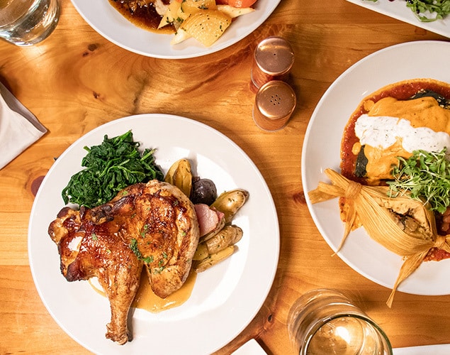 Three plates of food on a wooden table: grilled chicken with greens and potatoes, a tamale with sauce, and another dish partially visible. Nearby are salt and pepper shakers and a glass of water.