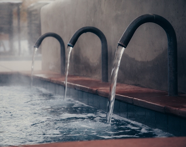 OSF Salt pool pipes winter Three curved metal pipes are pouring water into a steaming pool, creating a serene and relaxing atmosphere. The background has a soft, muted tone, enhancing the tranquil setting.