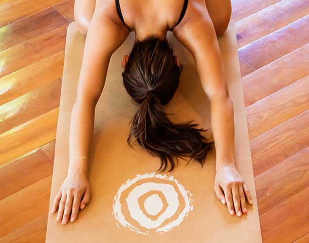 A person in a black sports bra performs a yoga child's pose on a brown mat with a white circular design. The floor is wooden, and the person's hair is tied back in a ponytail.