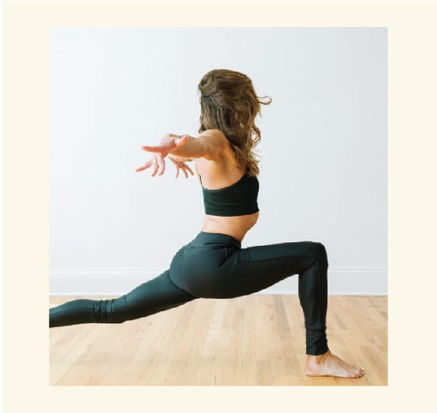 A person in athletic wear performs a yoga warrior pose on a wooden floor, facing sideways. The setting is minimal with a light, neutral background, emphasizing their balance and form.