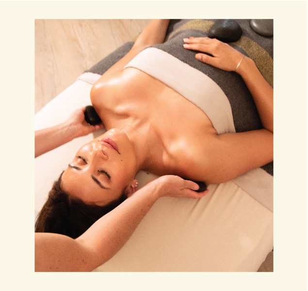 A person receiving a hot stone massage. They are lying on a massage table with eyes closed, covered by a towel. Smooth stones are placed on their body while a masseuse massages their shoulders. The setting is serene and calming.