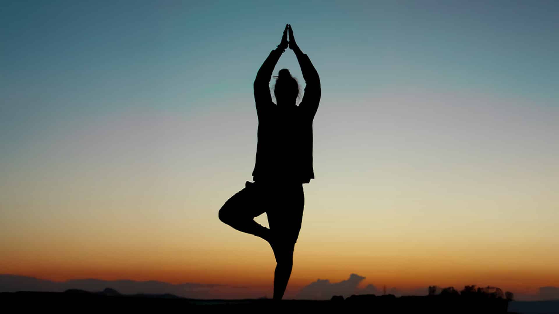 Silhouette of a person practicing a yoga pose outdoors at sunset. The individual stands on one leg with hands pressed together above their head, against a gradient sky transitioning from orange to blue.