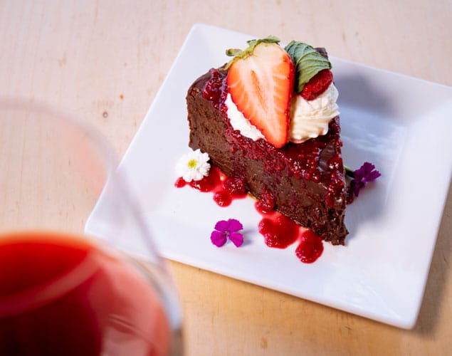 A slice of chocolate cake topped with whipped cream, a sliced strawberry, and red sauce sits on a square white plate. The cake is garnished with small flowers. In the foreground, a glass of red liquid is slightly out of focus.