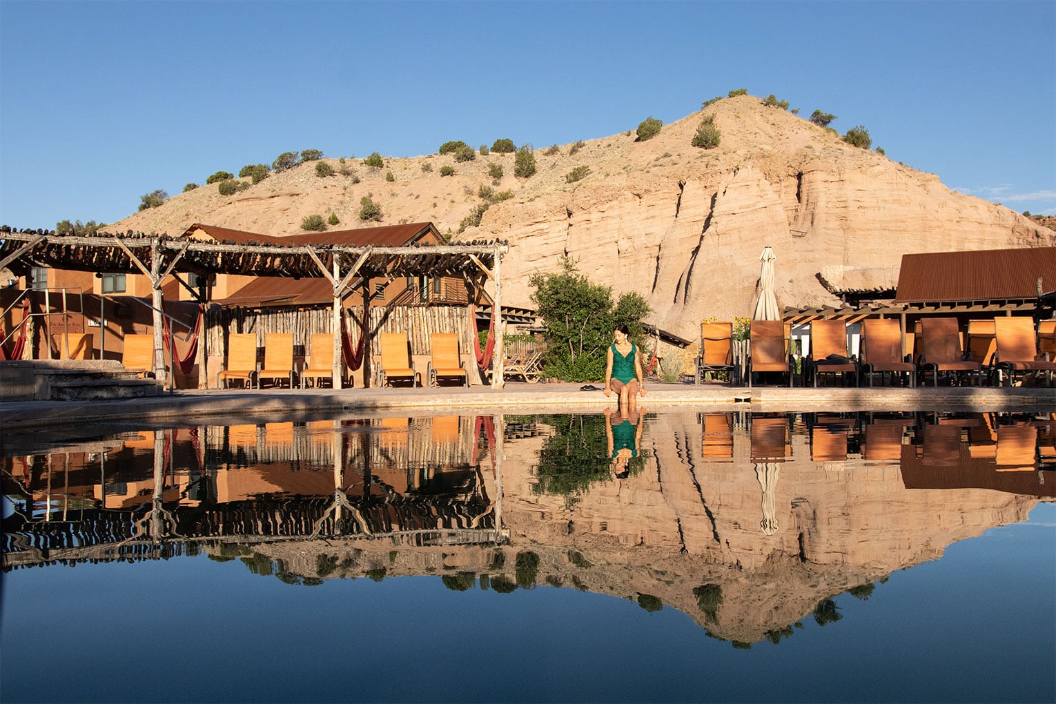 OC_SaltWaterPool_Cliffside_1530x1020 A person sits at the edge of a calm pool, with rustic wooden buildings and lounge chairs reflected in the water. A rocky hillside rises in the background under a clear blue sky.