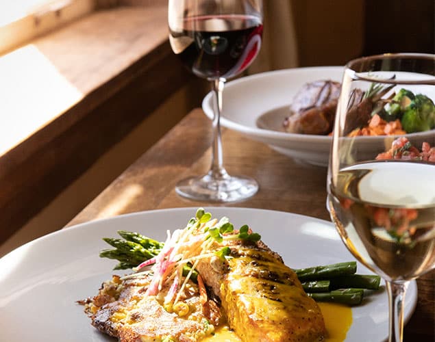 OC_Artesian_Dinner_Website_635x500 A plate with grilled fish, asparagus, and hash browns sits on a wooden table next to glasses of red and white wine, with sunlight streaming through a nearby window.