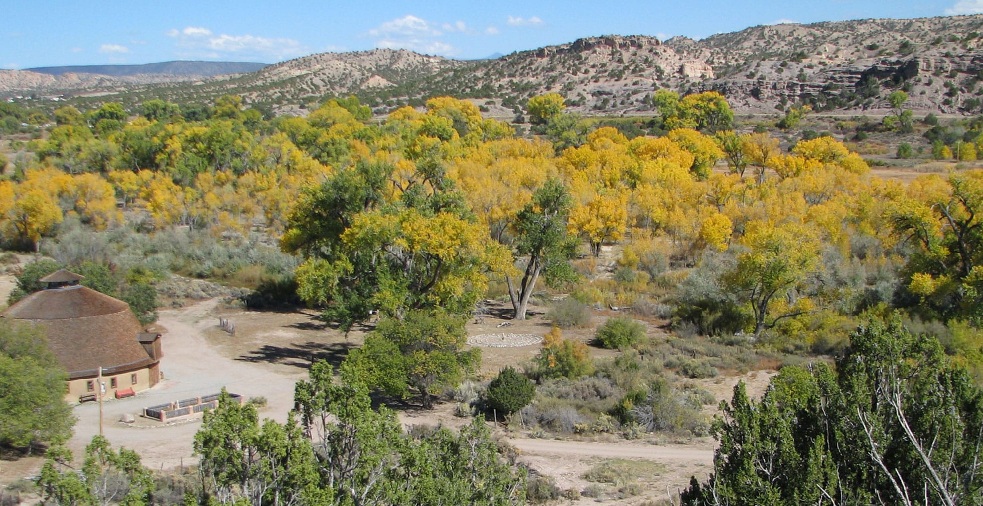 A landscape view of a valley with yellow-leaved trees, a small round building with a brown roof, dirt paths, and distant hills under a blue sky with scattered clouds.
