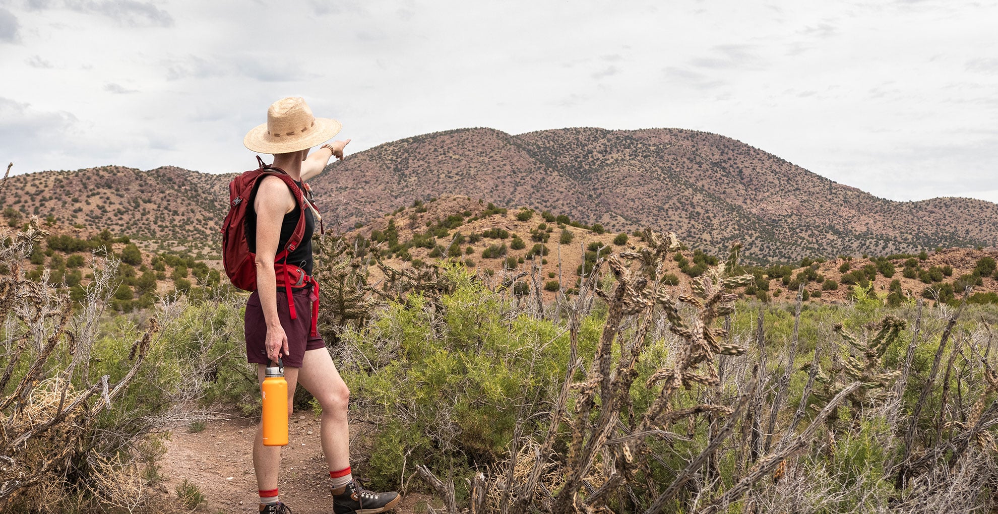 OC_Hike_Angie_Mountain_Website_1980x1020 A person wearing a hat, backpack, and hiking gear stands on a desert trail, holding a water bottle and pointing toward distant, brush-covered hills under a cloudy sky.