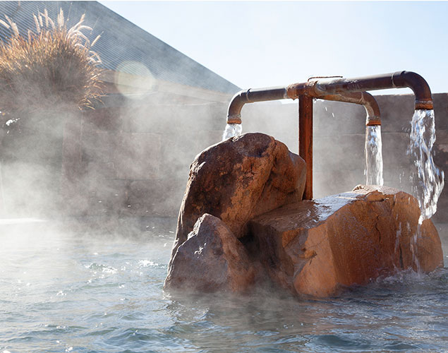 A close-up of hot spring water flowing from metal pipes over large rocks, with steam rising from the water. Sunlight shines on the scene, and a building with tall grass is visible in the background.