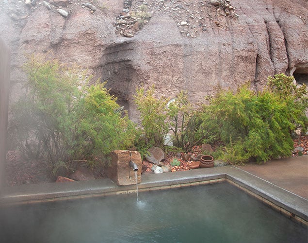 OC_Private-Pool_Faucet-With-Cliff_Website_635x500 A steaming outdoor hot spring pool with a small spout, surrounded by green shrubs and set against a rocky, natural landscape.