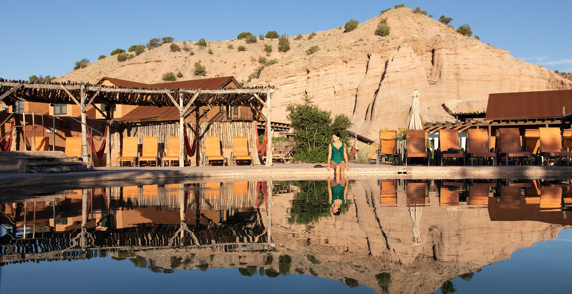 A person sits at the edge of a pool reflecting wooden lounge chairs, canopies, and sandstone cliffs in a desert resort setting under a clear sky.