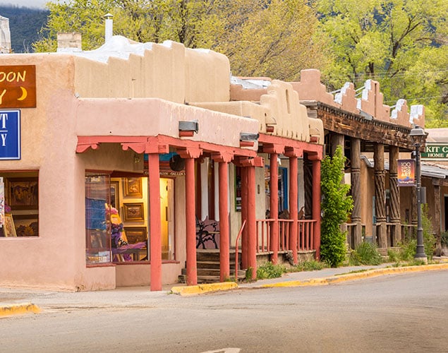 Row of adobe-style buildings with wooden beams and colorful pillars along a quiet street, featuring art galleries and greenery in the background, set in a small Southwestern town.