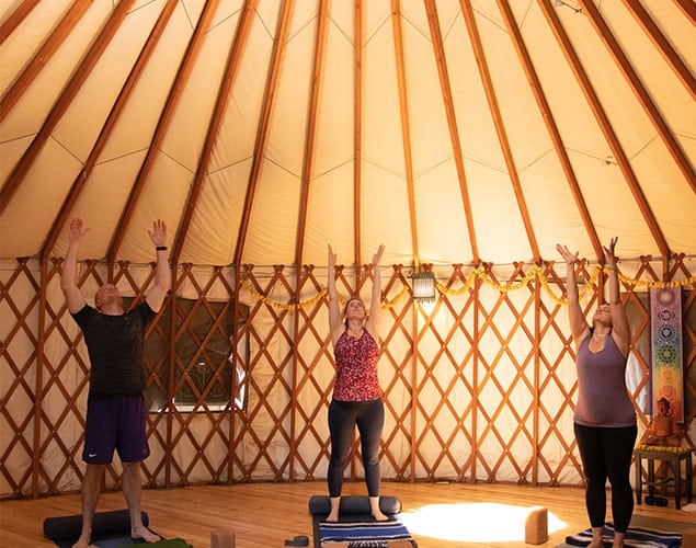 OC_Yoga_Yurt-3_Website_635x500 Three people stand with arms raised, practicing yoga inside a sunlit yurt with wooden lattice walls, yoga mats, and props on the floor. Warm light filters through the circular tent ceiling.