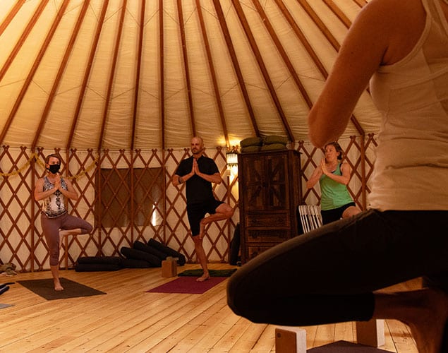 OC_Yoga_Yurt_Standing_Website_635x500 Four people practice yoga inside a round, wooden yurt. Three are in tree pose facing the instructor, who is pictured in the foreground. Soft lighting and yoga mats create a calm, focused atmosphere.