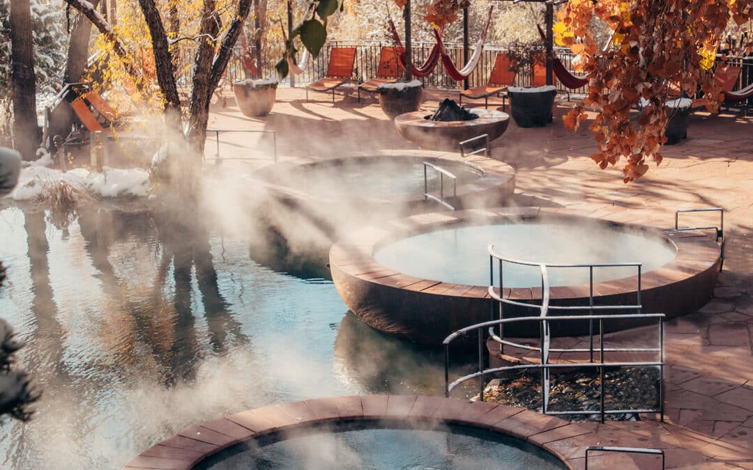Three round hot spring pools with steamy water outdoors, surrounded by trees with autumn leaves and some snow. Hammocks and chairs are in the background on a stone patio.