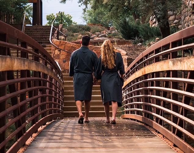 OSF_Spa_Robes-Couple-on-Bridge_Website_635x500 Two people wearing dark robes and sandals walk hand-in-hand across a wooden bridge toward stone steps, surrounded by trees and greenery in an outdoor setting.