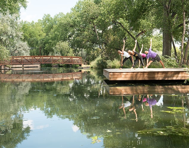 OSF_Yoga_Dock_Website_635x500 Three people practice yoga on a wooden platform beside a calm pond, surrounded by trees. Their reflections are visible in the water, and a red footbridge crosses the pond in the background.