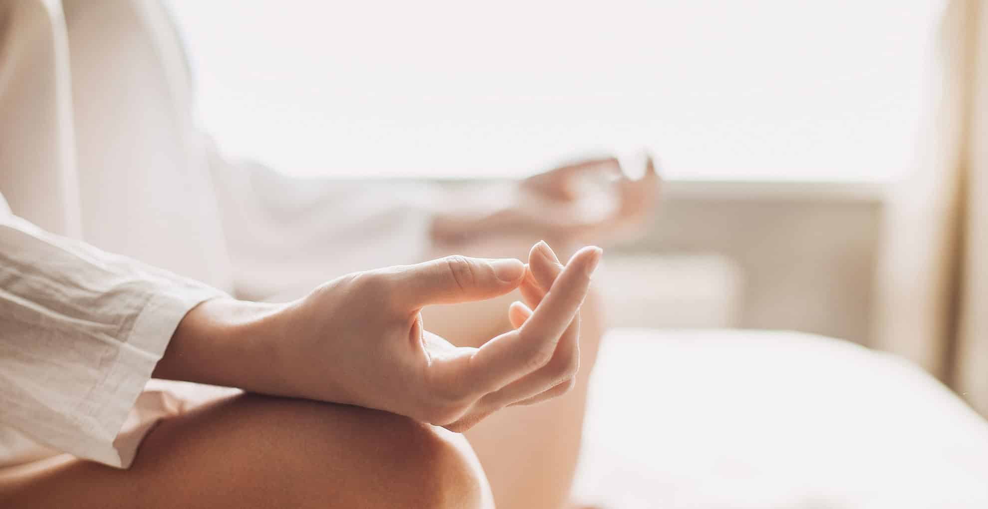 OSR_Meditation-Hands_Stock_Website_1980x1020 Close-up of a person sitting cross-legged on a bed, wearing a light robe, with hands resting on knees in a meditative pose, fingers gently touching, in a softly lit, peaceful room.