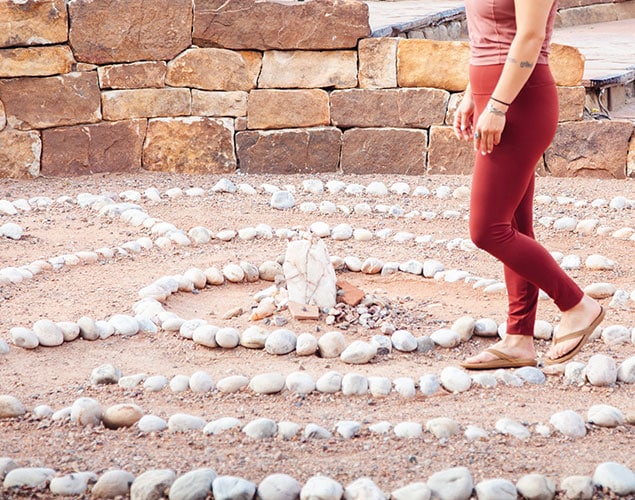 A person wearing maroon leggings and tan shoes walks through a stone labyrinth outdoors, with a central stone formation and a background of stacked stone walls.