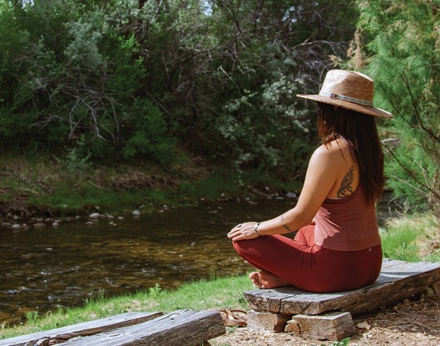 A woman wearing a hat and maroon clothes sits cross-legged on a wooden bench by a creek, surrounded by green trees, facing the water and nature.
