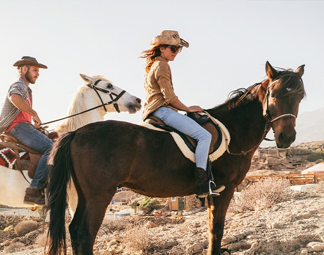 Two people wearing cowboy hats ride horses outdoors on a sunny day. The person in front is on a dark brown horse, while the person behind rides a white horse. Rocky terrain and mountains are visible in the background.