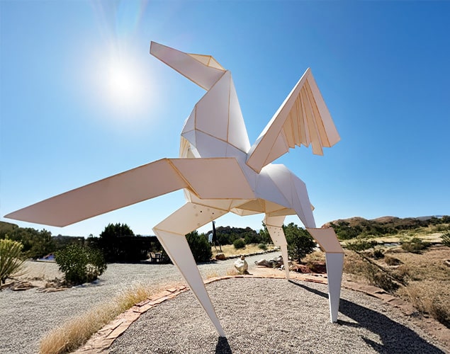 A large white sculpture resembling an origami pegasus stands outdoors on a gravel path under a bright blue sky with the sun shining. The landscape includes grass, bushes, and distant hills.