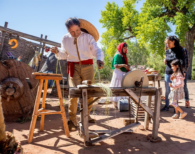 A man in historical clothing demonstrates traditional rope-making outdoors, using a wooden tool. Nearby, women and children watch, some dressed in period costumes. The scene is sunny with trees and rustic equipment in the background.