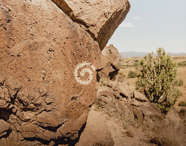 A large brown rock with a white spiral petroglyph stands in the foreground; sparse vegetation and a tree are in the background under a clear sky.