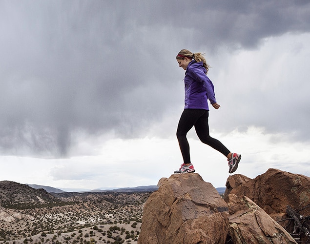 A woman in athletic clothing stands on a rocky peak, appearing to jump or step down, with a dramatic cloudy sky and expansive desert landscape in the background.