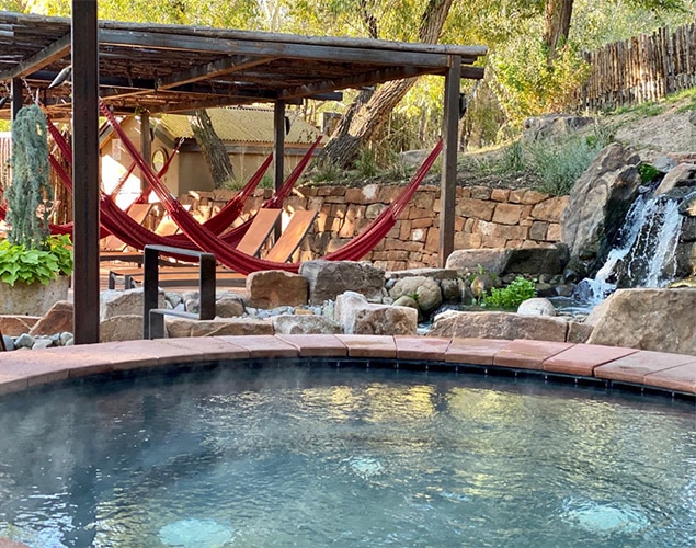 A round hot tub with steam rising, set in a natural outdoor setting. Behind it are red hammocks hanging under a pergola, stone walls, rocks, green plants, and a small waterfall flowing into a pond.