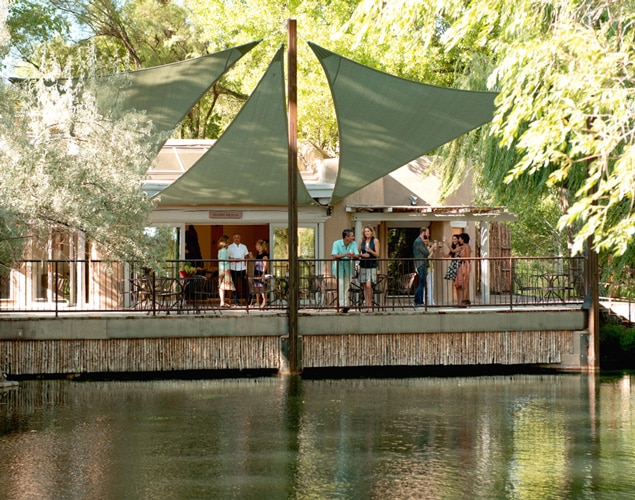 OjoSantaFeSpaResortMoonHouse A group of people socialize on a shaded outdoor patio overlooking a pond, surrounded by trees and greenery, under large triangular fabric canopies.