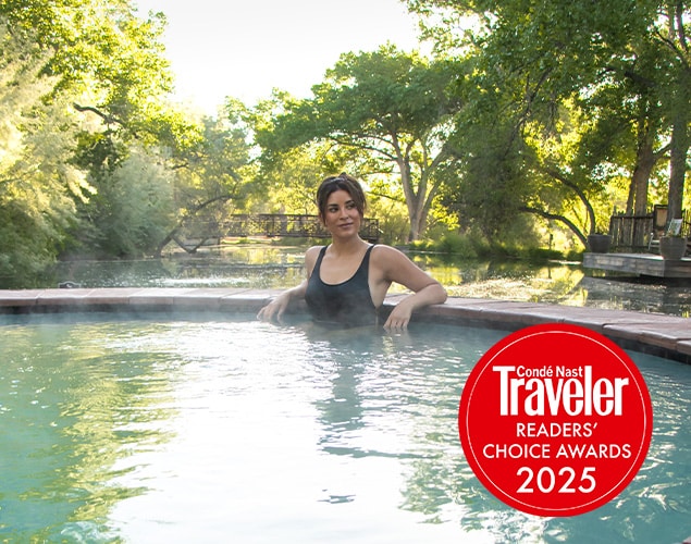 A person in a black swimsuit relaxes in an outdoor hot spring surrounded by lush greenery, with a "Condé Nast Traveler Readers' Choice Awards 2025" badge in the lower right corner.