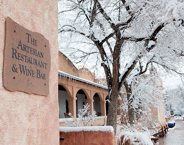 635x500_Artesian_Snow A sign for The Artesian Restaurant & Wine Bar is mounted on a stucco wall, with snow-covered trees and adobe-style buildings visible in the background.