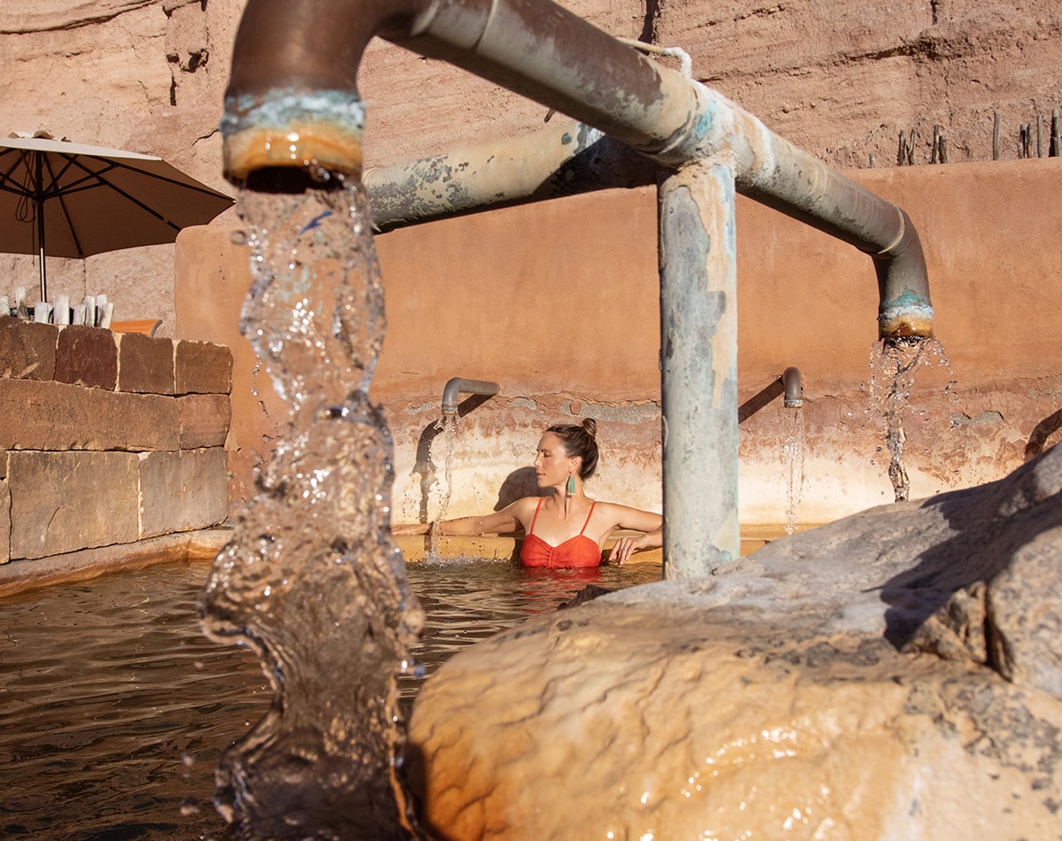 A woman in a red swimsuit relaxes in a natural hot spring pool, with water flowing from metal pipes, surrounded by stone walls and desert rocks under a sunny sky.