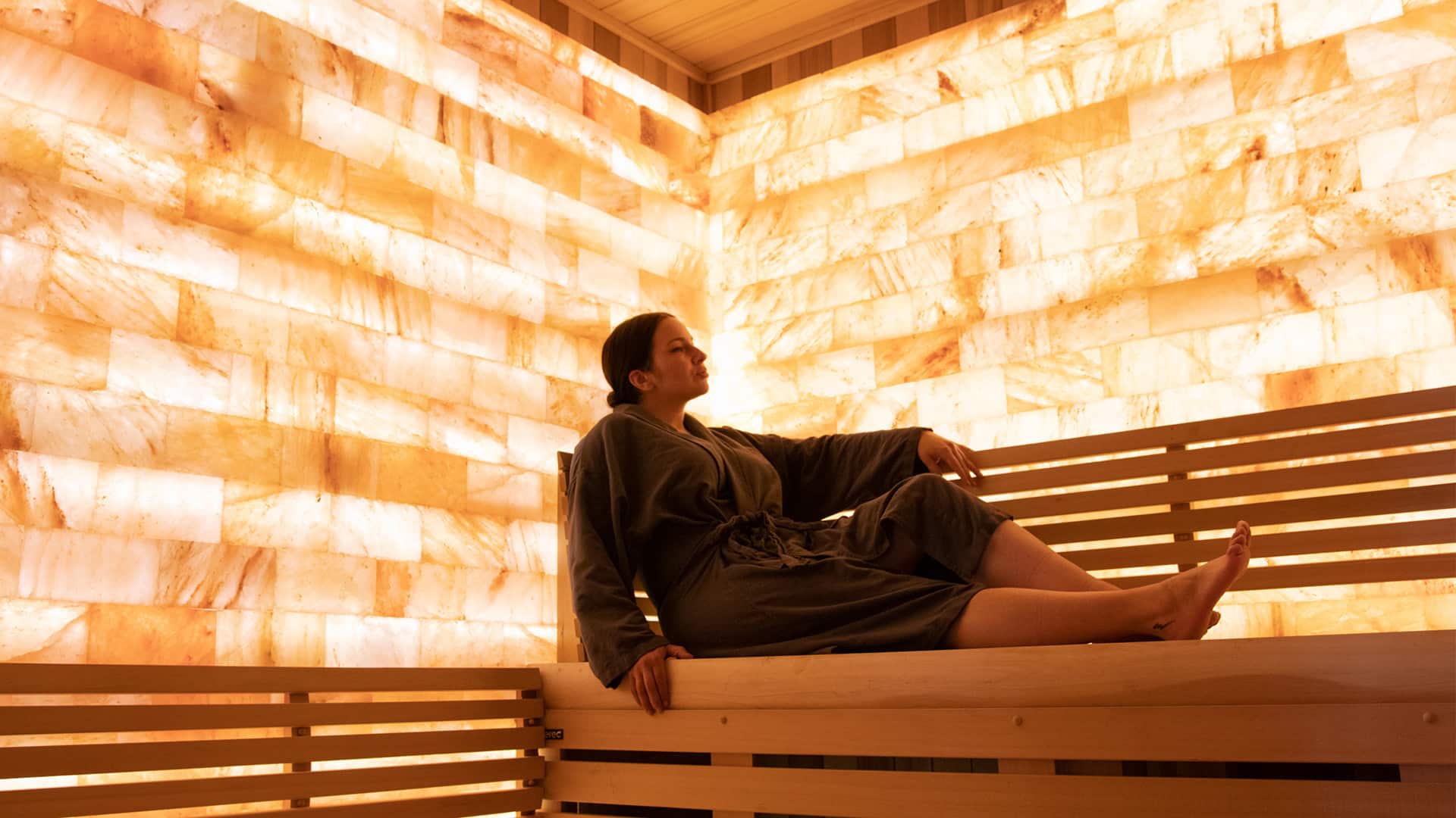 1920x1080_OC_SAUNA A person in a dark robe relaxes on a wooden bench inside a sauna with warmly lit, glowing salt block walls.