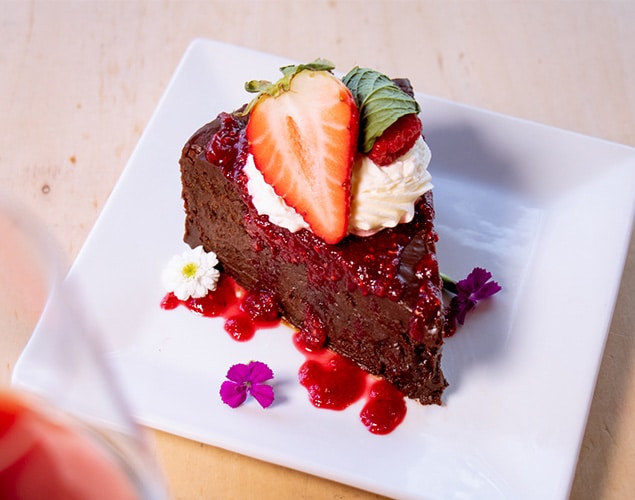 A slice of chocolate cake topped with whipped cream, a strawberry half, raspberry sauce, and small edible flowers, served on a white square plate.